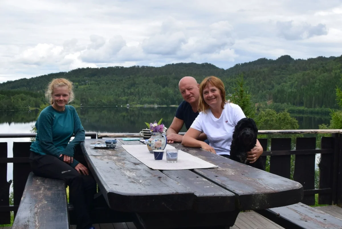 Tre personer sitter rundt et bord ute på terassen, med vannkanten like bak, foto