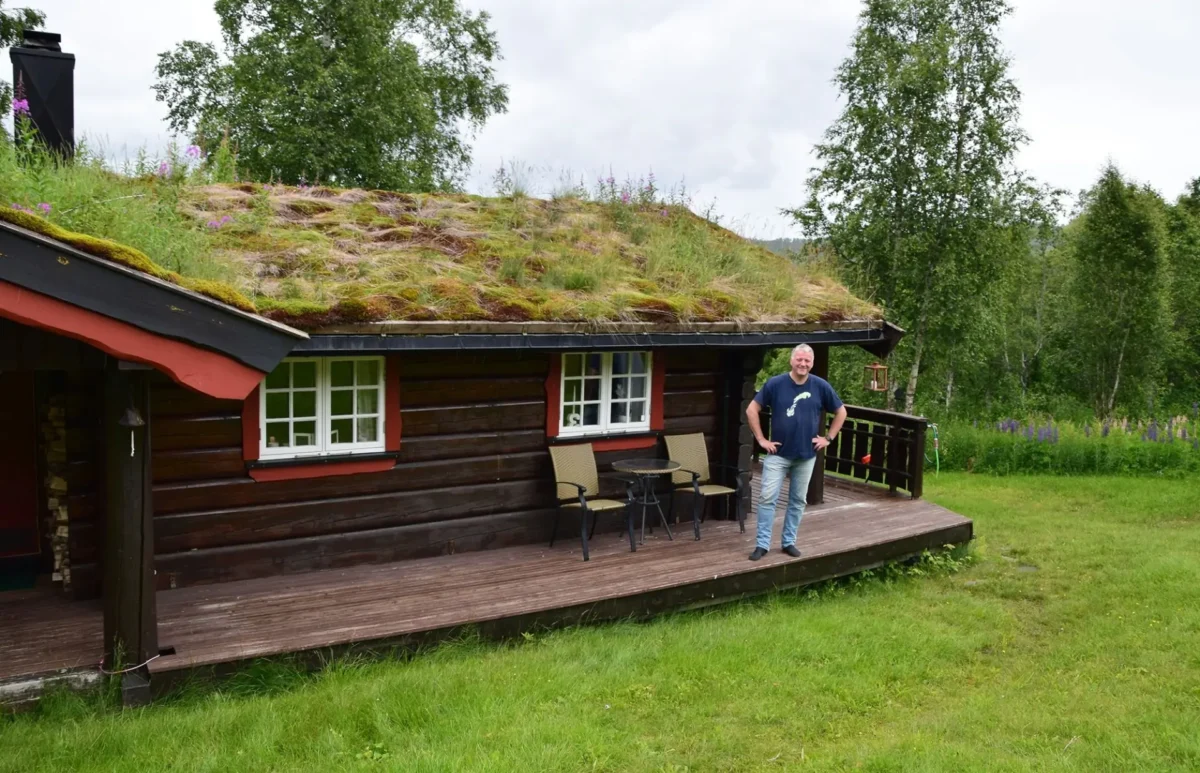 Mann står på terassen på tømmerhytte med torvtak, foto