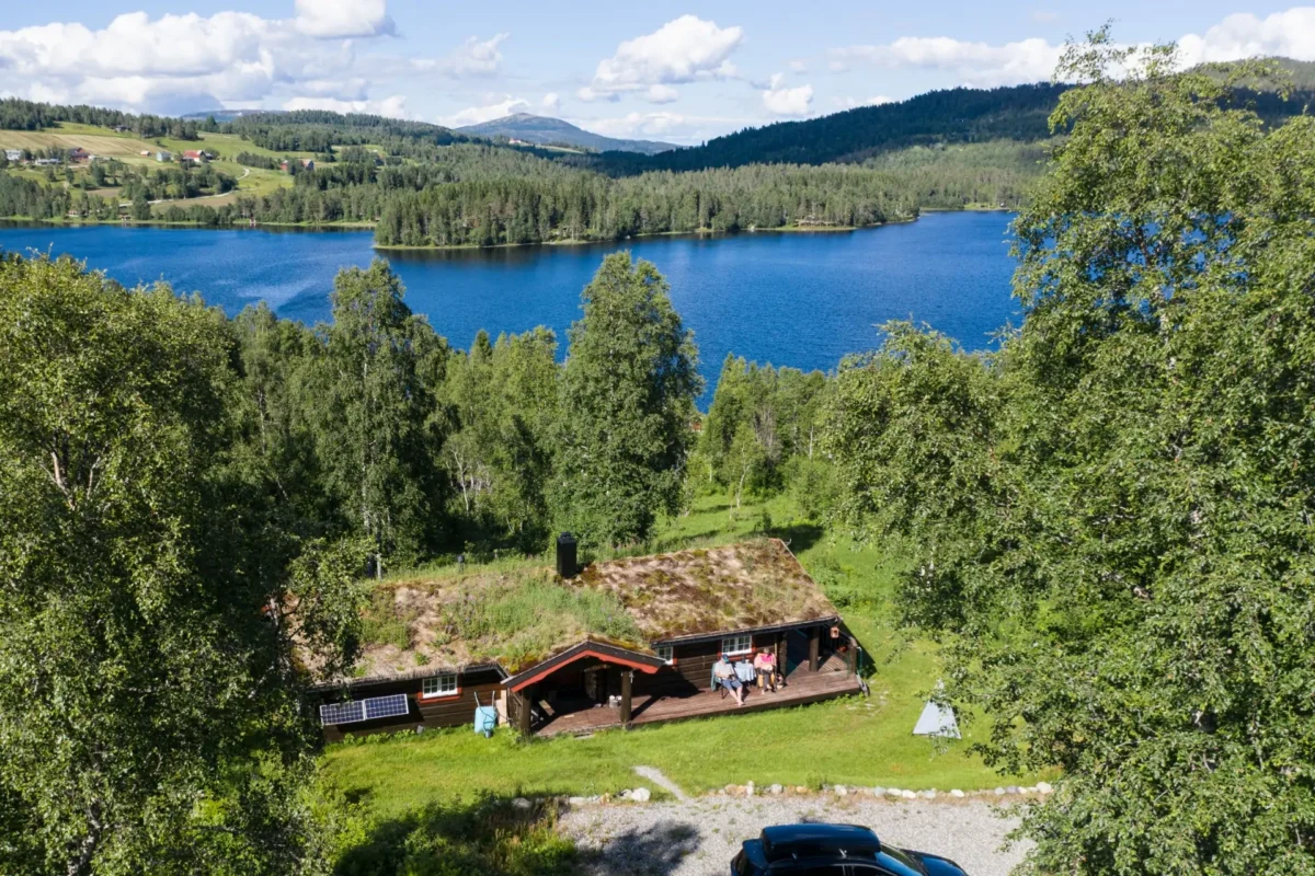 Hytte med trær rundt, vann og fjell bak, sommerlig, dronefoto