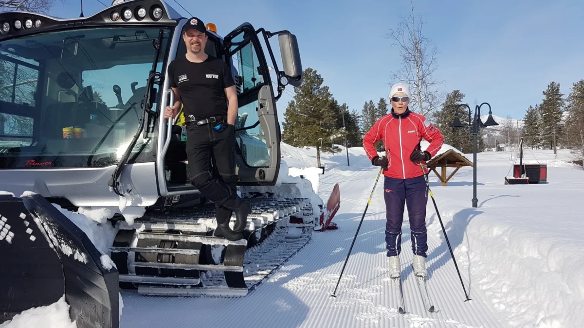 En person står i døra på ei løypemaskin og en skigåer står ved siden av, foto