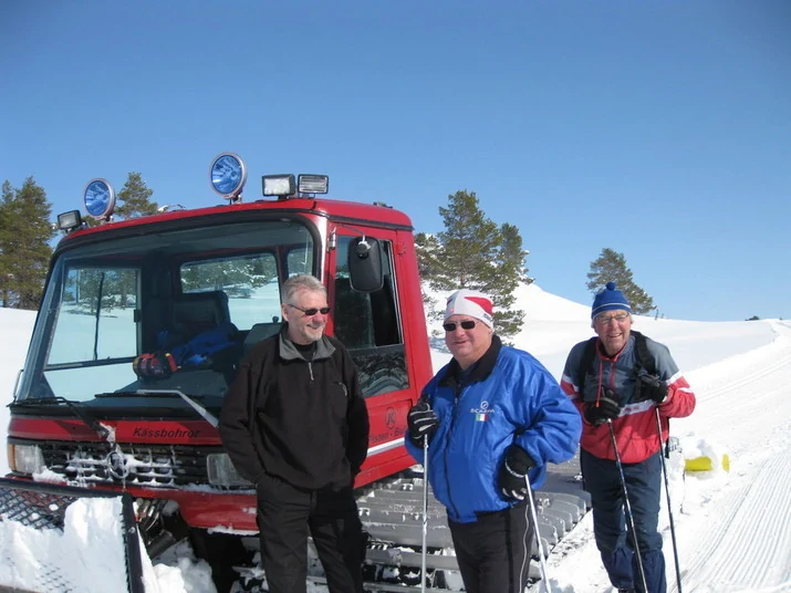 Tre personer foran en løypemaskin, to av de går på ski, foto