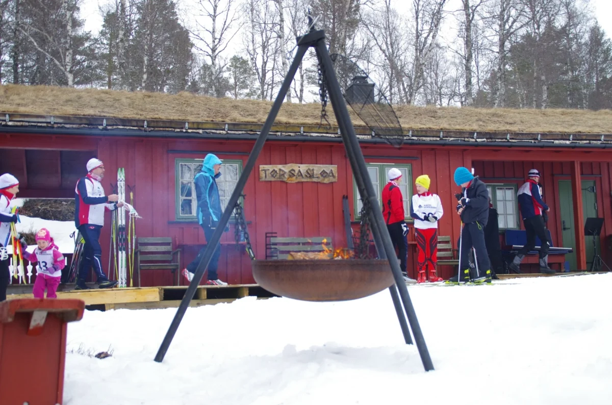 Bålpanne foran trebygg, skigåere i bakgrunnen, foto