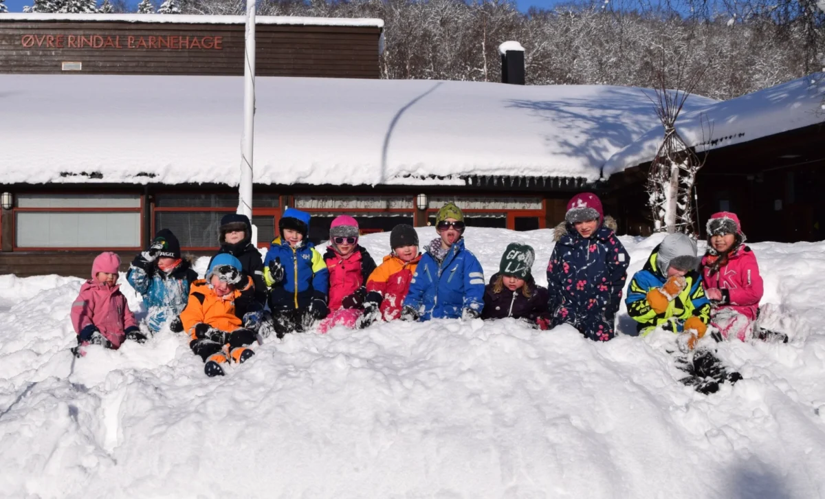 Barnehagebarn sitter på rekke i snøen, foto