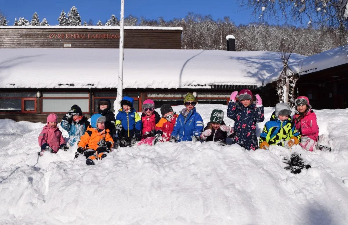 Barnehagebarn sitter på rekke i snøen, foto